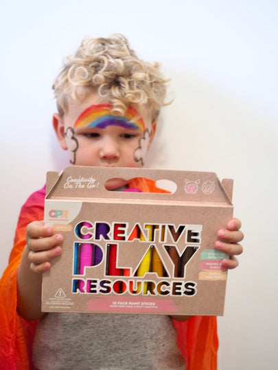 Child holding a box labeled 'Creative Play Resources' with rainbow face paint.