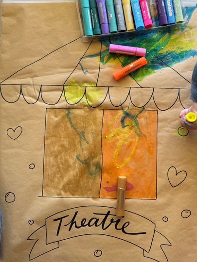 Children's cardboard theater with colorful markers and a child's hand holding a marker.