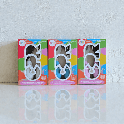 Three colorful boxes of children's cutlery sets on a white surface with a textured wall background.