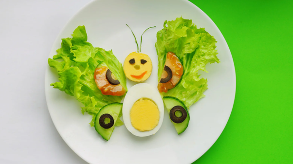 A creative butterfly-shaped food arrangement on a white plate, made with boiled eggs, lettuce, cucumber, olives, and salmon, designed to make healthy eating fun for kids.