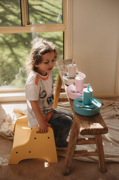 Child sitting on yellow stool with colorful silicone dinnerware plates on a wooden table, enjoying mealtime play.