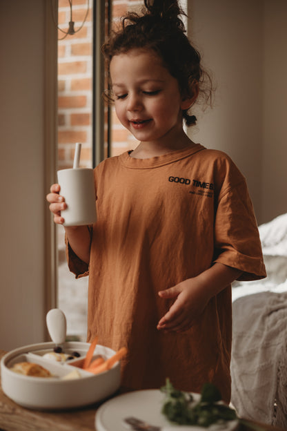 Child in a brown shirt with 'Good Times' text, holding a cup in a casual indoor setting.