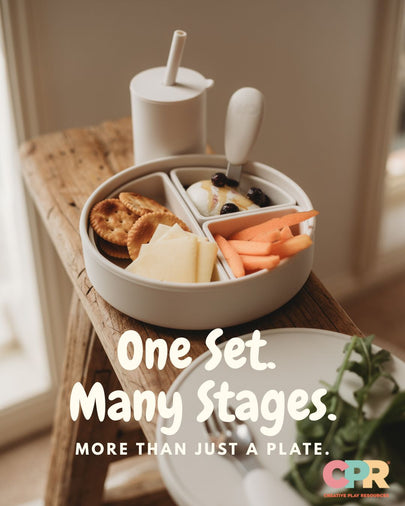 A styled breakfast scene showing a round compartment plate set on a wooden table, filled with crackers, cheese, fruit, and a small cup with a straw. Large overlaid text reads 'One Set.