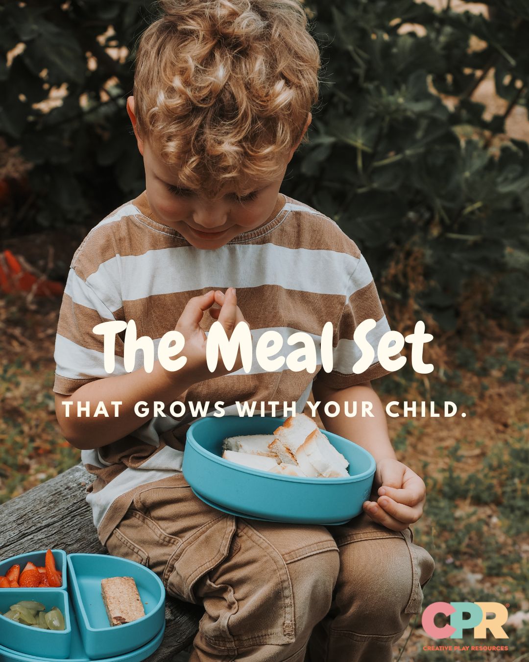 A young child sitting outdoors on a log, holding a bright blue divided lunch bowl with sandwich halves inside. Another blue lunch tray with snacks is visible in the lower left.