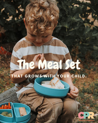 A young child sitting outdoors on a log, holding a bright blue divided lunch bowl with sandwich halves inside. Another blue lunch tray with snacks is visible in the lower left.
