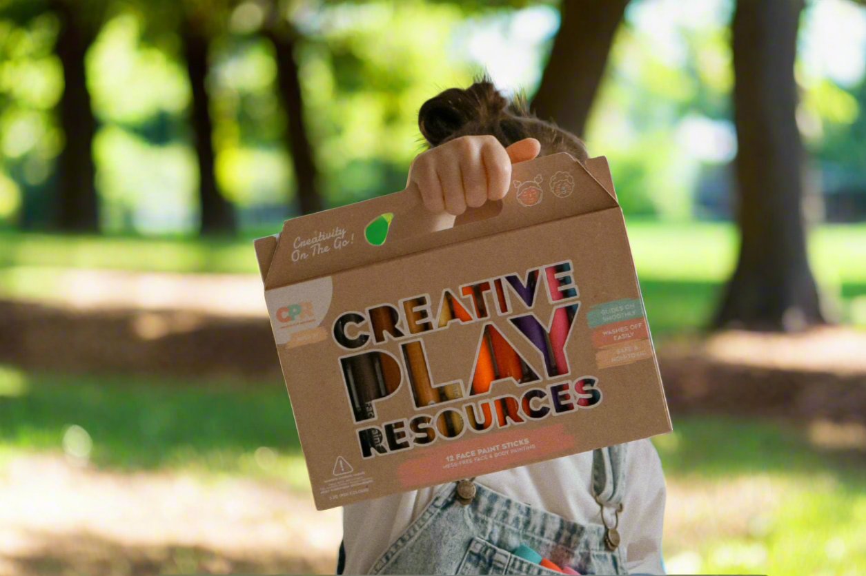 child outdoors holding a unique box of various coloured face paint sticks that twist up and down like a crayon.