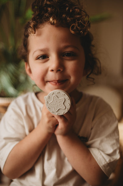 A smiling young child with curly brown hair holds a round, textured off-white soap or shampoo bar in front of their face, wearing a white t-shirt in a warm, softly blurred indoor setting.